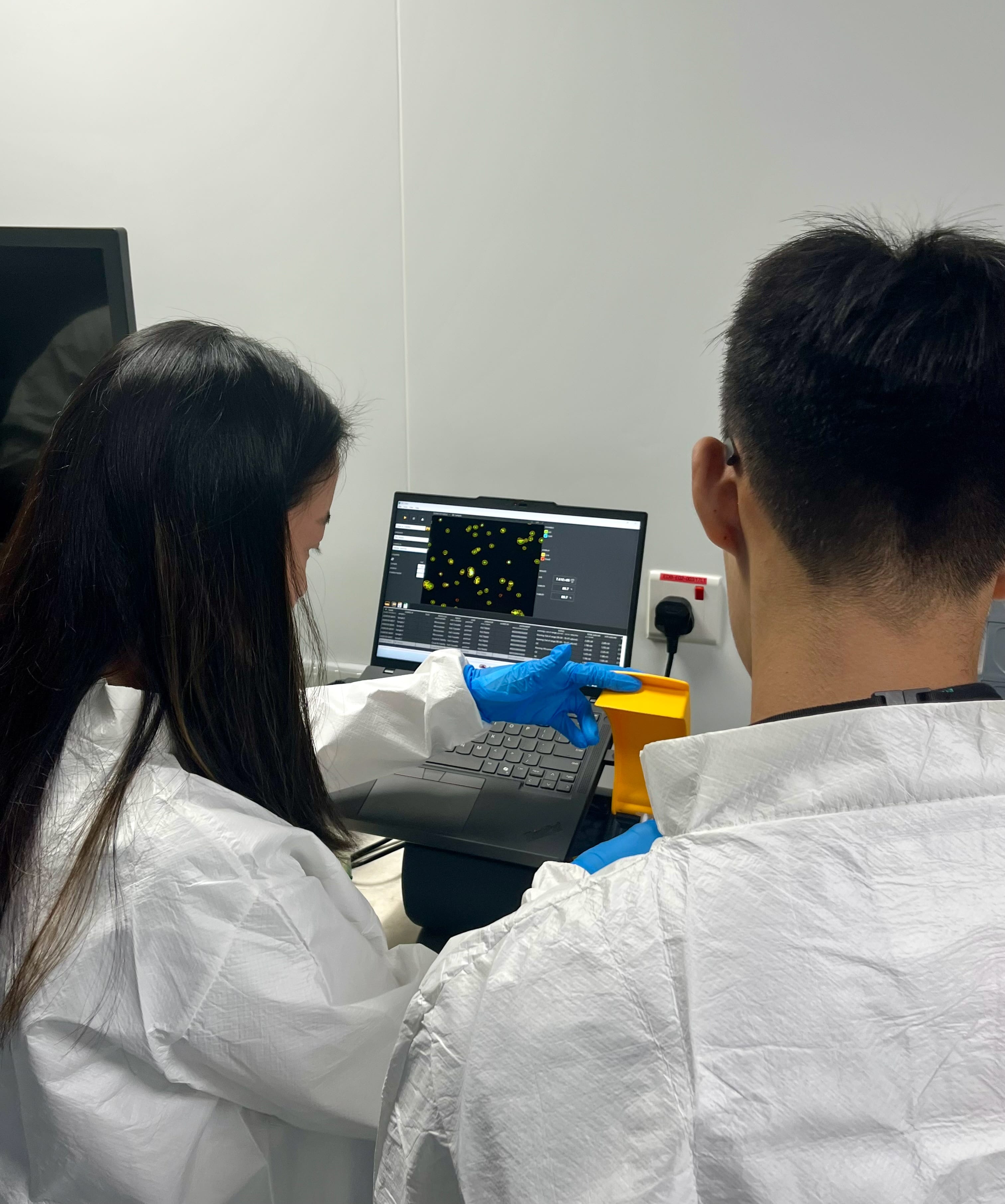 Participants in laboratory protective attire reviewing cell analysis results on a laptop during a hands-on cell culture training session in a research laboratory.